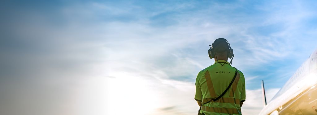 A ground operations employee seen from behind wearing a high‑visibility yellow Delta shirt and a headset, standing outdoors and facing the airfield under a blue sky.