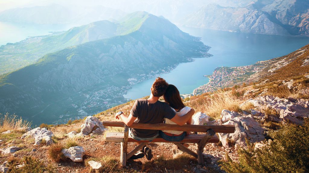 Two people sit side by side on a wooden bench overlooking a coastal landscape, with a blue body of water and a town visible below and mountains rising in the distance under clear daylight.