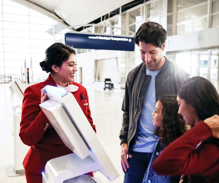 A Delta employee in a red uniform assists a family of three at a self‑service kiosk in a bright, modern airport concourse.