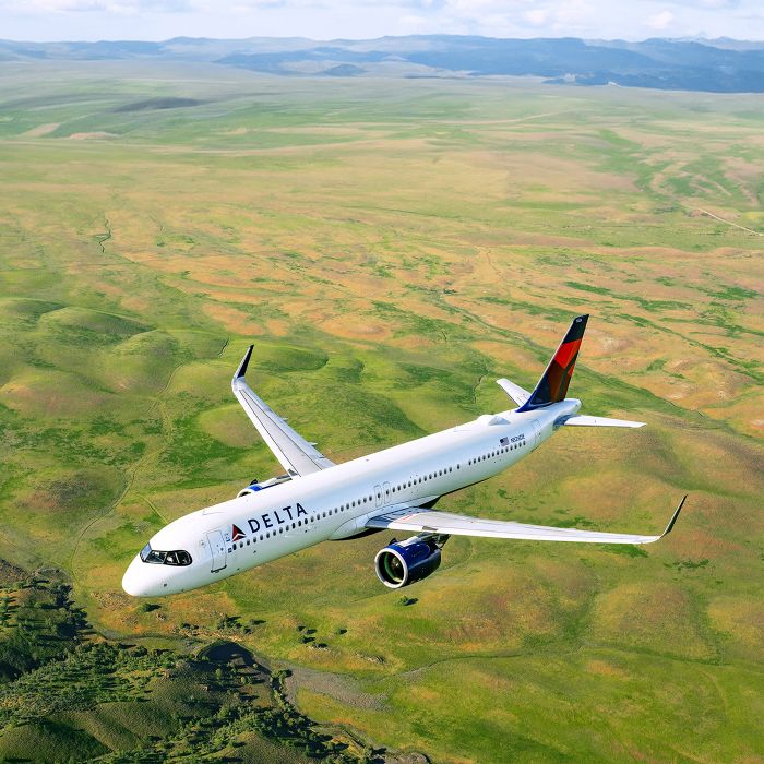 A Delta airplane flies above a green landscape of fields and rolling terrain, viewed from an elevated angle, with the aircraft’s white body and red‑and‑blue tail clearly visible