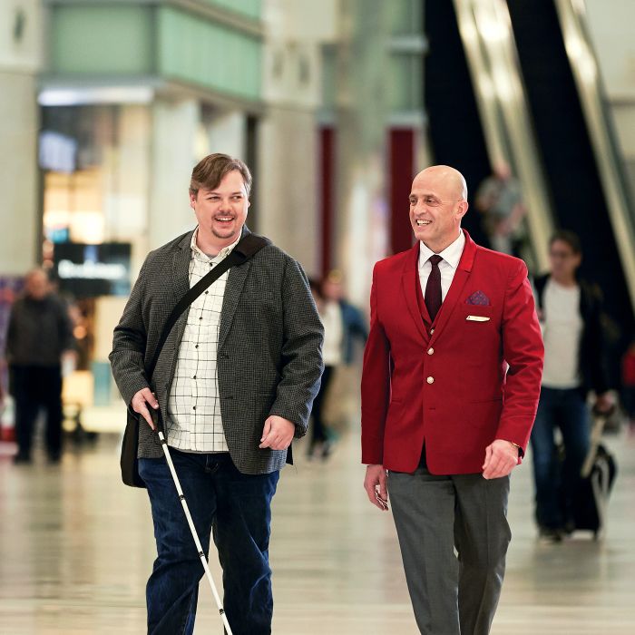 A Delta employee in uniform walks alongside a traveler using a white cane through the airport concourse, with other travelers visible in the background.