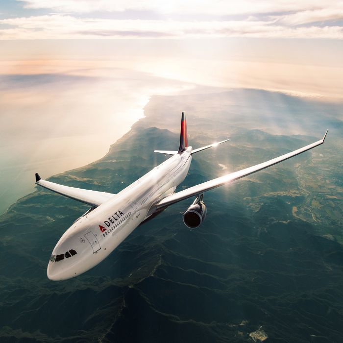 A Delta airplane viewed from above flies under a hazy sky above a rugged coastline and mountainous landscape. Sunlight reflects off the aircraft’s wings.