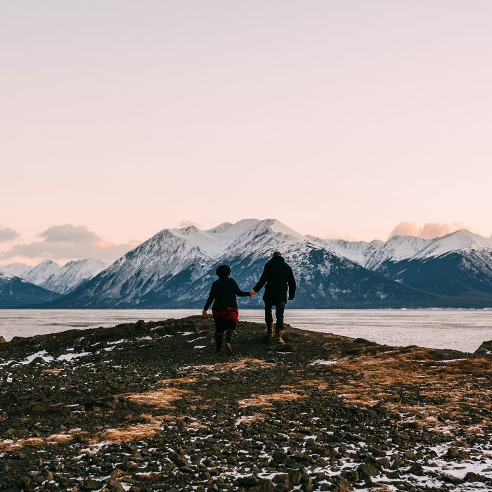 Two people walk hand in hand along a rocky shoreline, looking across a lake toward snow‑capped mountains under a soft, pastel sky.
