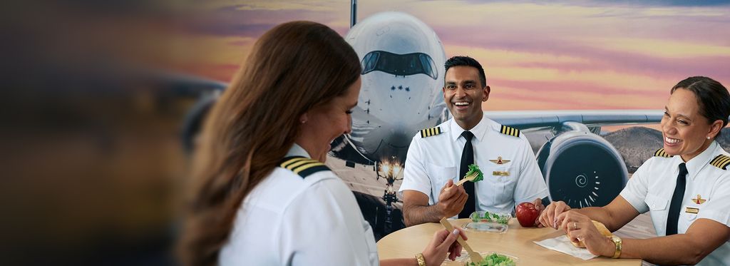 Three uniformed pilots sit at a table eating fresh fruit and salads, in front of a mural of a forward facing airplane and a colorful sunset sky.