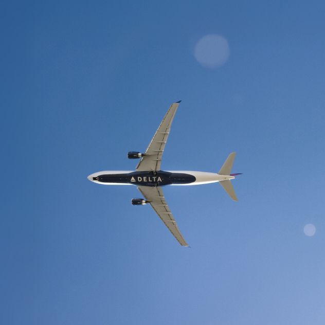 A Delta airplane flying across a clear blue sky, shown from an underside view. The word “DELTA” appears in large, white lettering on the bottom of the aircraft against the dark blue background.