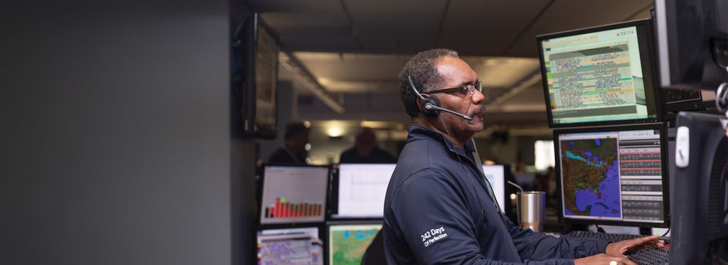A Delta employee wearing a headset stands at a workstation in an operations control center, facing multiple computer monitors displaying maps and flight‑tracking data