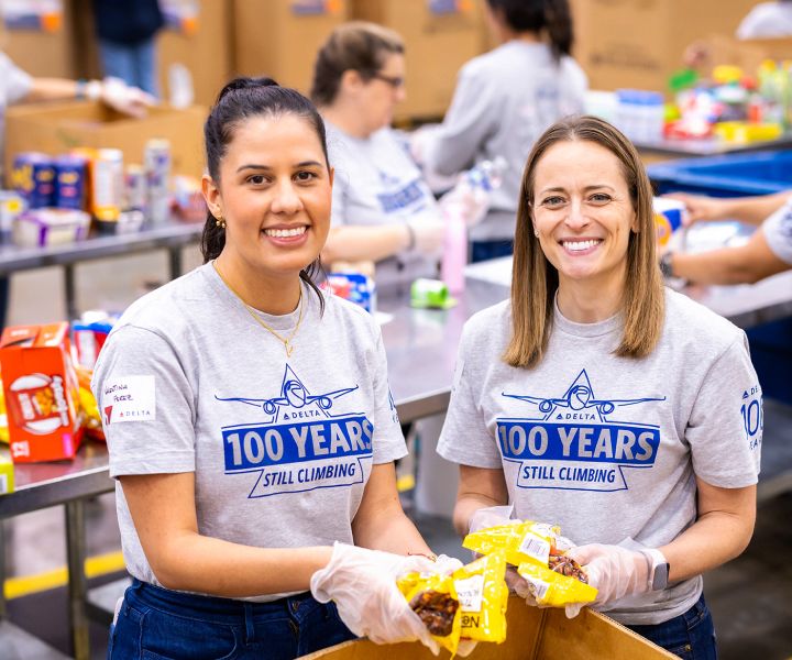 Two Delta employees stand side by side wearing matching gray T‑shirts that read “100 YEARS – STILL CLIMBING.” They hold packaged food items, with tables, boxes, and other volunteers working in the background