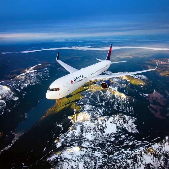 A Delta airplane flies above a river and snow‑dusted landscape, viewed from the front and slightly above. Patches of forest, water, and rugged terrain stretch into the distance beneath a partly cloudy sky