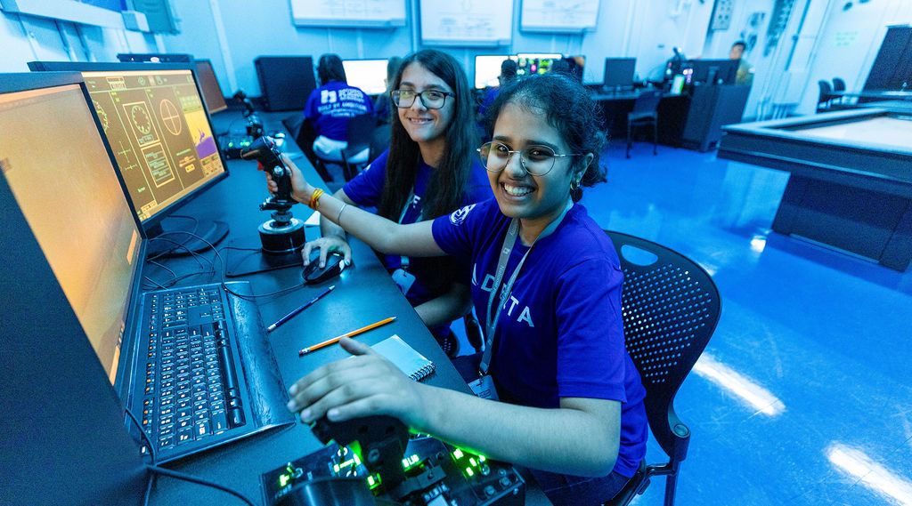 Two young people in blue Delta t-shirts sit at a workstation in a blue‑lit operations room, using computer consoles with large monitors displaying data and system interfaces. One holds a mouse while the other holds a joystick.