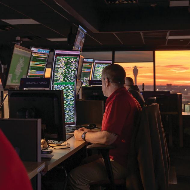 A Delta employee sits at a workstation in an operations control center, facing multiple computer monitors filled with data and maps. Large windows in the background reveal a sunset sky and airport structures in the distance.