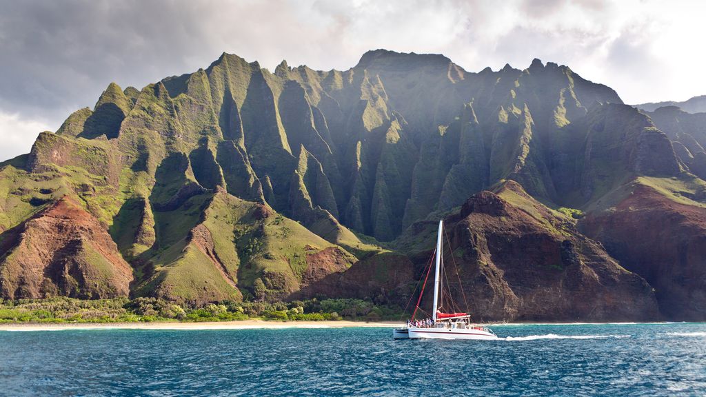 A sailboat moves across deep blue water near steep, green coastal cliffs, with rugged mountains rising sharply from the shoreline under a partly cloudy sky.