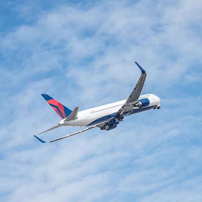 A Delta airplane climbs into a partly cloudy blue sky, viewed from below with its wings extended and landing gear visible as it departs.
