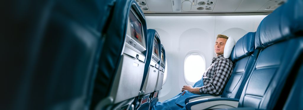 Inside an airplane cabin, a passenger wearing a black-and-white checkered shirt rests in a blue leather window seat with eyes closed, head on a white pillow and legs covered with a blue blanket, as daylight comes through the window