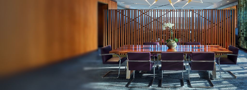 A modern conference room with a long wooden table surrounded by purple upholstered chairs. A potted plant sits at the center of the table, with vertical wood slats and glass walls in the background.