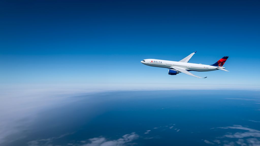 A Delta airplane flies across a clear blue sky in side profile, with its white fuselage, blue engines, and red‑and‑blue tail logo clearly visible.