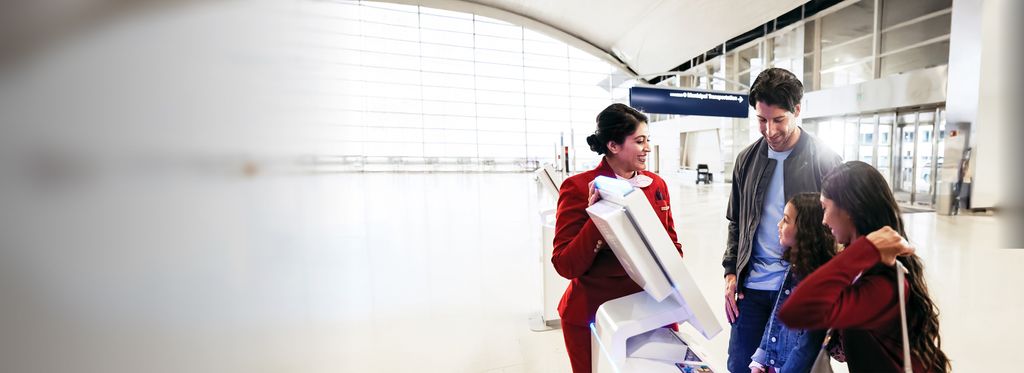 A Delta employee in a red uniform assists a family of three at a self‑service kiosk in a bright, modern airport concourse.