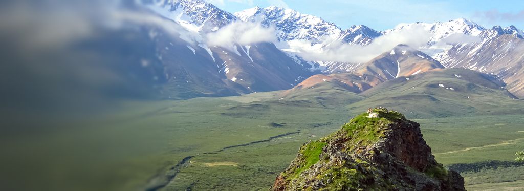 A rugged mountain landscape with snow‑capped peaks in the distance and rolling green valleys below. In the foreground, a rocky outcrop rises above the surrounding terrain under a partly cloudy sky.