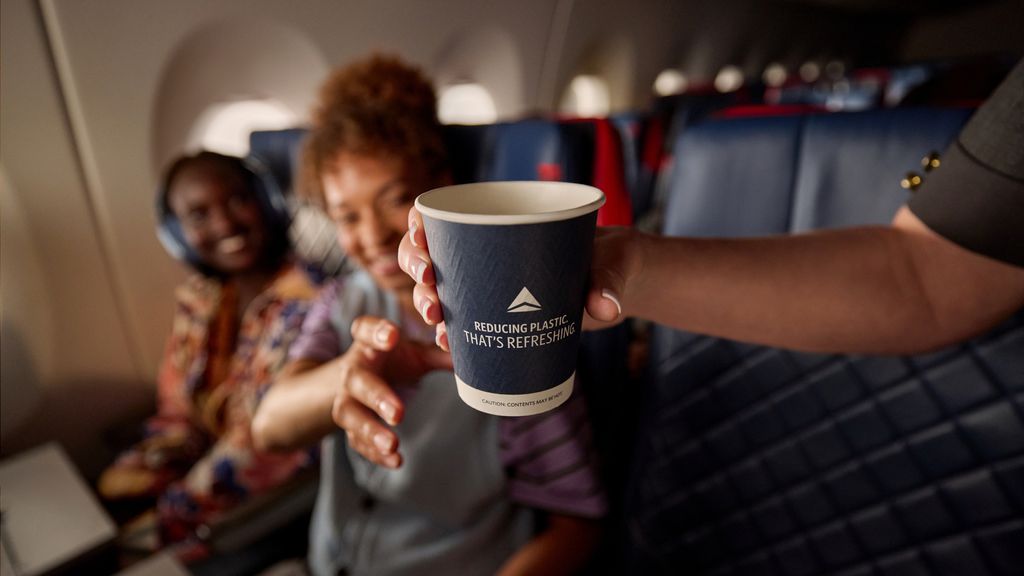 Inside an airplane cabin, a flight attendant’s hand offers a dark blue paper cup to a seated passenger. The cup reads, “Reducing plastic. That’s refreshing.” Another passengers sit in blue leather seat in the background.