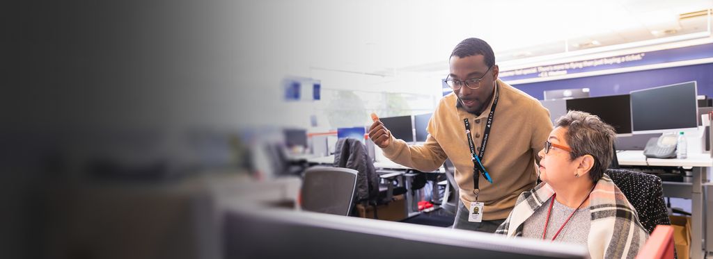 Two office employees collaborate at a workstation. One stands behind the desk looking at a computer monitor, while the other sits at the desk. Rows of desks, monitors, and office equipment fill the background.
