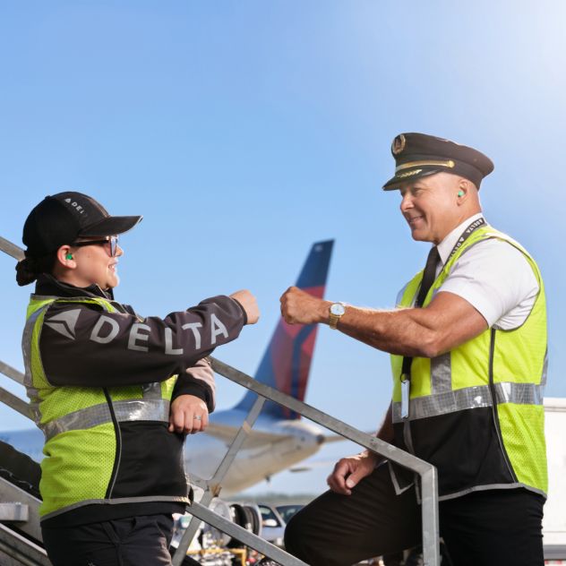 A Delta pilot and mechanic wearing high‑visibility vests exchange a fist bump outdoors at the aircraft boarding stairs beside an airplane under a clear blue sky.
