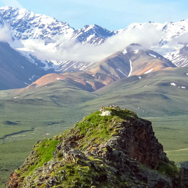 A rugged mountain landscape with snow‑capped peaks in the distance and rolling green valleys below. In the foreground, a rocky outcrop rises above the surrounding terrain under a partly cloudy sky.
