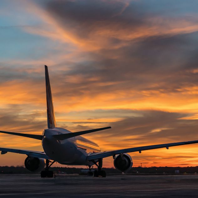 A Delta airplane is seen from behind on a tarmac, silhouetted against a dramatic orange and gold sunset sky.