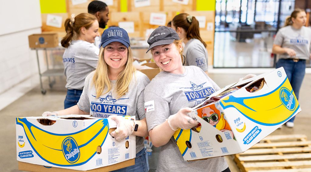 Two Delta volunteers wearing matching gray “100 Years – Still Climbing” T‑shirts and gloves work together to pack boxed items at a table, with other volunteers and supplies visible in the background