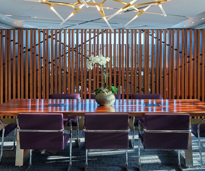 A modern conference room with a long wooden table surrounded by purple upholstered chairs. A potted plant sits at the center of the table, with vertical wood slats and glass walls in the background.