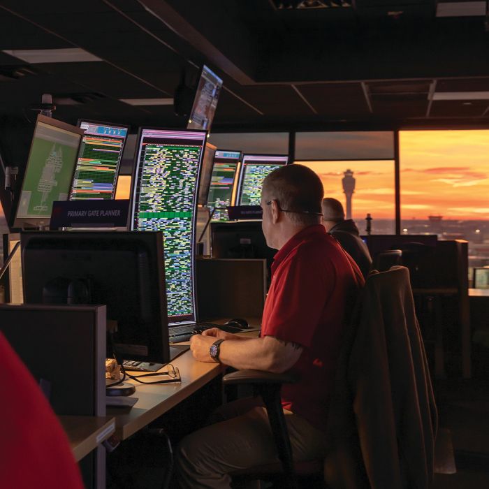 A Delta employee sits at a workstation in an operations control center, facing multiple computer monitors filled with data and maps. Large windows in the background reveal a sunset sky and airport structures in the distance.
