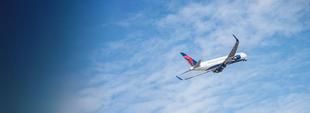 A Delta airplane climbs into a partly cloudy blue sky, viewed from below with its wings extended and landing gear visible as it departs.