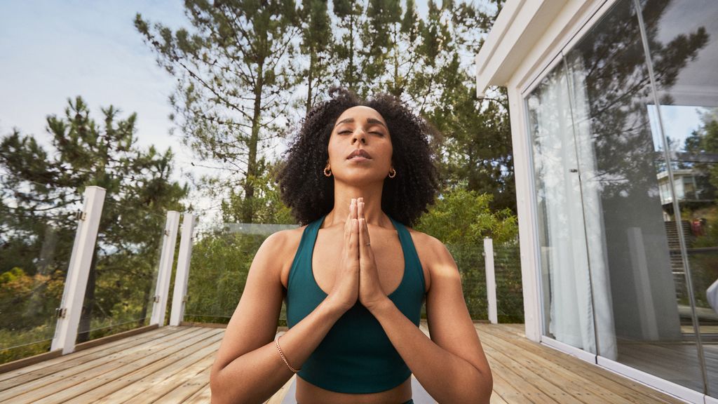 A person stands outdoors with hands pressed together at chest level in a prayer or meditation pose. They are wearing a sleeveless green top, with trees and greenery visible in the background.