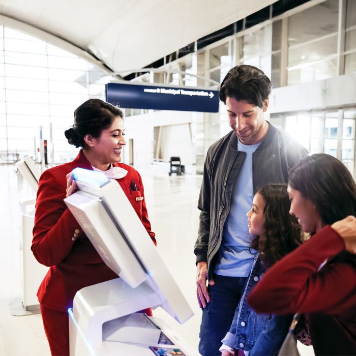 A Delta employee in a red uniform assists a family of three at a self‑service kiosk in a bright, modern airport concourse.