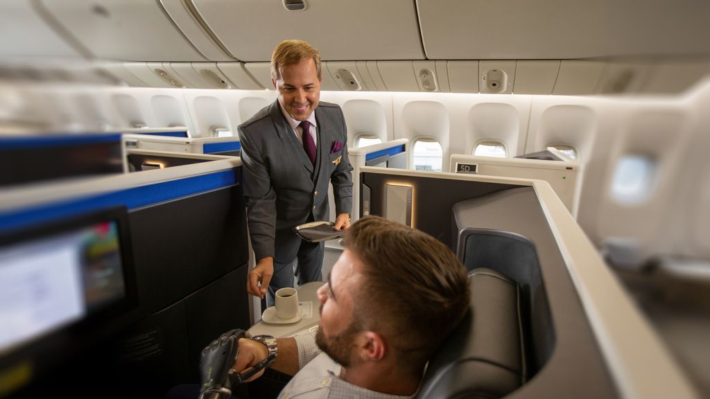 Inside a premium airplane cabin, a uniformed flight attendant stands beside a seat, holding a tray and offering a drink to a seated passenger with a robotic hand. Individual suites with privacy dividers, overhead bins, and soft cabin lighting are visible in the background.