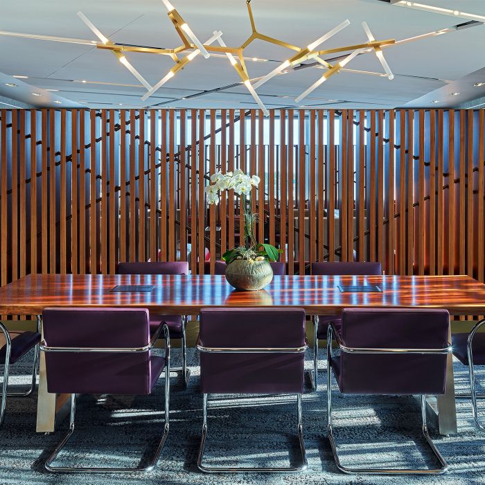 A modern conference room with a long wooden table surrounded by purple upholstered chairs. A potted plant sits at the center of the table, with vertical wood slats and glass walls in the background.