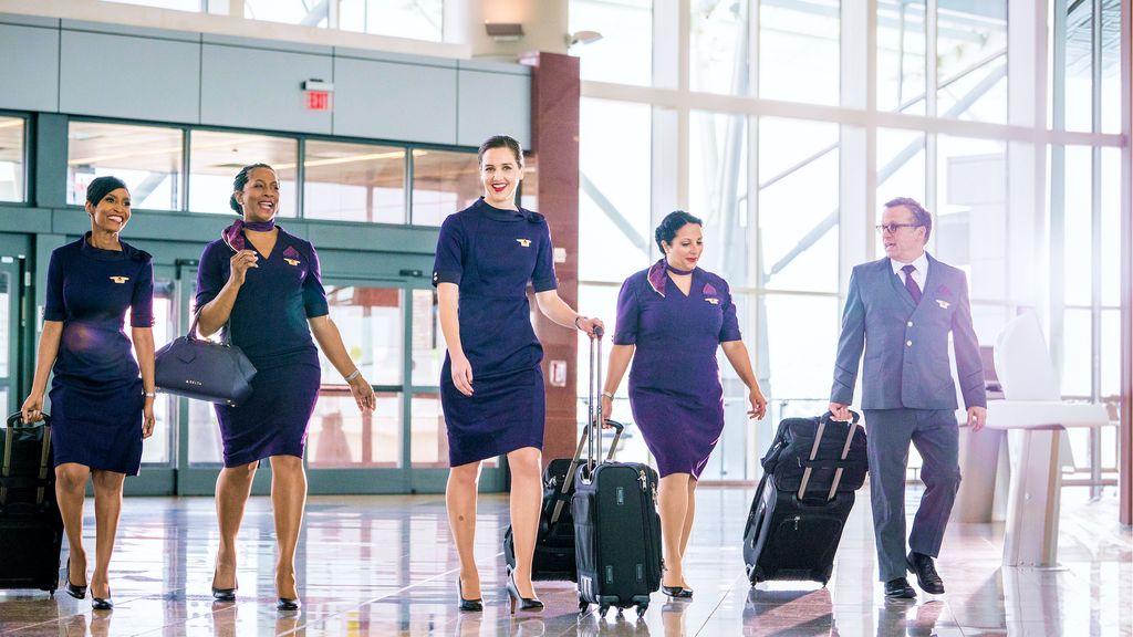 Delta employees in uniform walk together through a bright airport terminal, pulling rolling suitcases, with large windows and natural light visible in the background.