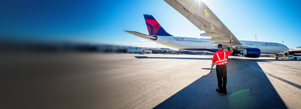 An airport ramp scene shows a ground crew member wearing an orange high‑visibility jacket standing on the tarmac and signaling with a handheld baton to a Delta airplane, assisting it as it parks at the gate under a clear blue sky.