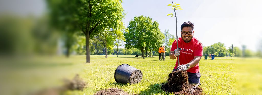 A Delta employee volunteer wearing a red “Delta in the Community” T‑shirt and work gloves kneels on grass while holding a plant, preparing to place it in the ground. Other volunteers and trees are visible in the background.