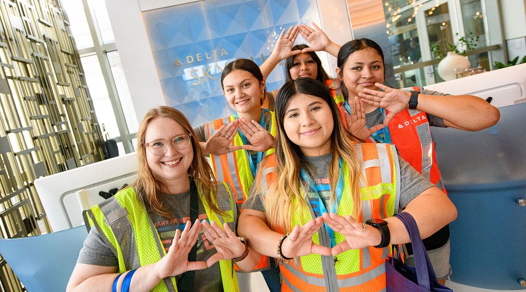 A group of young people wearing high-visibility safety vests stands together indoors, forming triangle shapes with their hands in front of their chests. A Delta Sky Club sign and glass walls are visible in the background