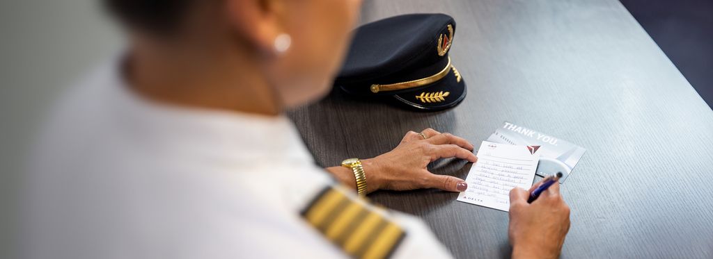 Close-up view of a uniformed Delta pilot seated at a desk, writing a handwritten note on a Delta-branded “Thank You” card. A pilot’s hat rests on the desk beside the card. The pilot’s sleeve with striped insignia is visible, emphasizing a professional aviation setting and a gesture of appreciation.