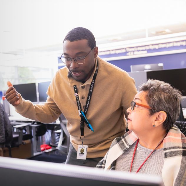 Two office employees collaborate at a workstation. One stands behind the desk looking at a computer monitor, while the other sits at the desk. Rows of desks, monitors, and office equipment fill the background.