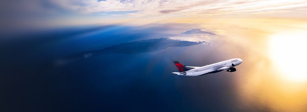 A Delta airplane flies above a sunlit ocean at golden hour, viewed from the side with warm light reflecting off the fuselage and wings against a glowing sky.