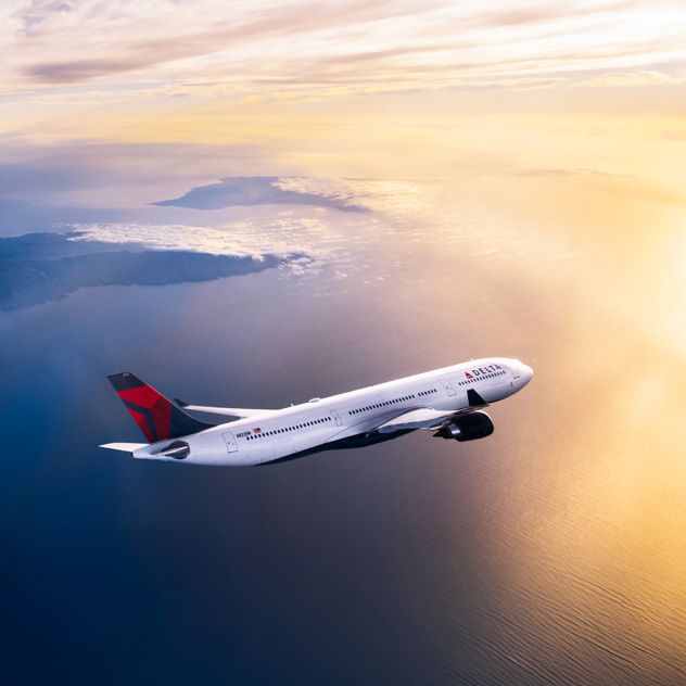 A Delta airplane flies above a sunlit ocean at golden hour, viewed from the side with warm light reflecting off the fuselage and wings against a glowing sky.