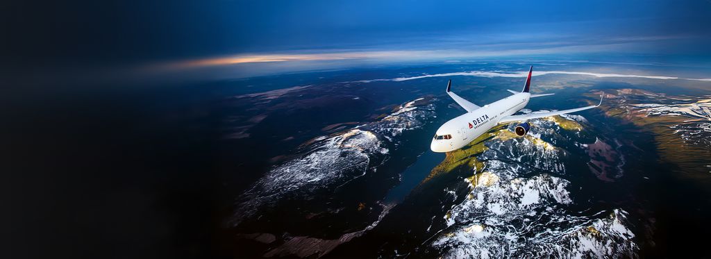 A Delta airplane flies above a river and snow‑dusted landscape, viewed from the front and slightly above. Patches of forest, water, and rugged terrain stretch into the distance beneath a partly cloudy sky