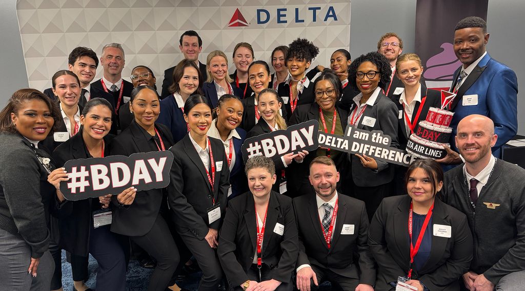 A group of Delta employees poses together indoors in front of a wall with the Delta logo. They are wearing coordinated dark attire with name badges, and red lanyards. Several people hold black signs reading “#BDAY” and “I am the Delta Difference”