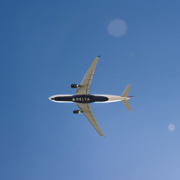 A Delta airplane flying across a clear blue sky, shown from an underside view. The word “DELTA” appears in large, white lettering on the bottom of the aircraft against the dark blue background.