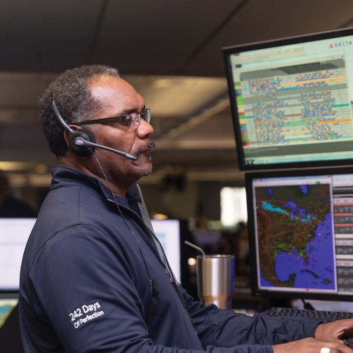 A Delta employee wearing a headset stands at a workstation in an operations control center, facing multiple computer monitors displaying maps and flight‑tracking data
