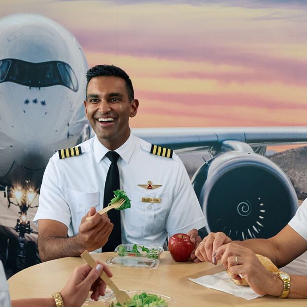 Three uniformed pilots sit at a table eating fresh fruit and salads, in front of a mural of a forward facing airplane and a colorful sunset sky.