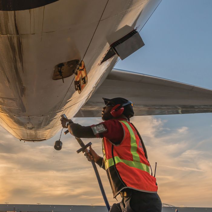 A Delta ground operations employee in an orange high-visibility vest holds a hose that connects to the underside of a Delta aircraft on the tarmac at sunset.