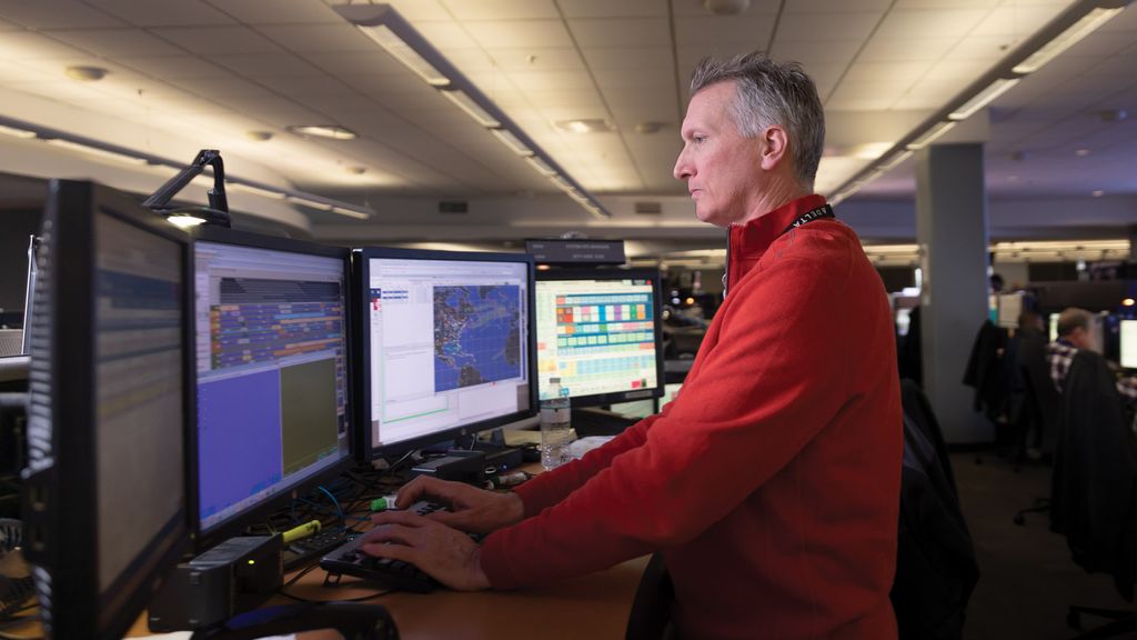 An operations control center employee in a red pullover stands at a computer workstation with multiple large monitors displaying maps and data. Other employees work at similar stations in the background under bright overhead lighting.