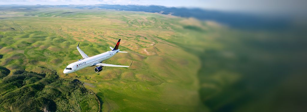 A Delta airplane flies above a green landscape of fields and rolling terrain, viewed from an elevated angle, with the aircraft’s white body and red‑and‑blue tail clearly visible